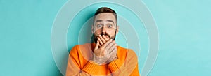 Close-up of shocked caucasian man covering his lips and mouth, staring astounded at camera, standing against turquoise