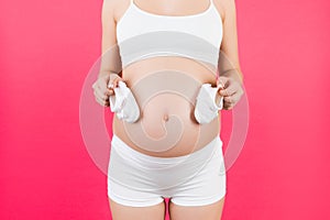 Close up of shoes for baby girl in pregnant woman`s hands at pink background. Future mother is wearing white underwear. Motherhoo