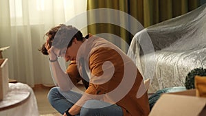 Close up shot of apartment living room. Man sitting on the floor holding head, shocked upset expression after receiving