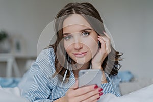 Close up shot of attractive brunette young woman downloads songs from network, uses smart phone for entertainment, dressed in nigh