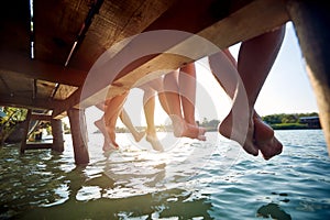 Close-up shot of a barefoot group of friends sitting on the dock on the river. Summer, river, vacation