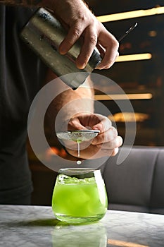 Close up shot of bartender hand pouring basil cocktail in a glass from shaker. Barman preparing classic modern basil