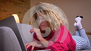 Close-up shot of blonde housewife in pink sweater lying on stomach on sofa surfing in laptop in cozy home atmosphere.