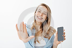 Close-up shot of carefree happy european girlfriend with fair hair, showing smartphone and pulling hand towards camera