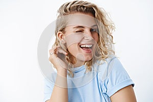 Close-up shot of coquettish and flirty cute blonde woman with short curly haircut winking and putting hair strand behind