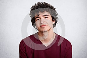 Close up shot of curly haired European teenager with little beard, wears maroon shirt. Young smiling male stands with lowered