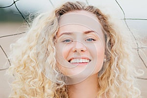 Close up shot of curly lovely young female has broad shining smile, looks positively at camera, has joyful expression, stands outd