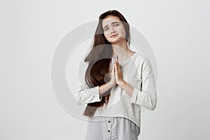 Close up shot of cute young hopeful dark-haired girl wearing casual long sleeved top pressing palms together, feeling