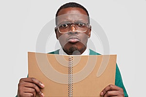 Close up shot of displeased young African American man with gloomy facial expression, purses lips, holds spiral notepad