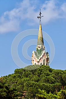 Close-up shot of Gaudi`s building roof top tower along green trees against blue sky in Park Guell, Barcelona, Catalonia