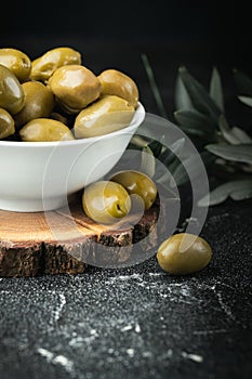 Close up shot of green olives in a white bowl on the wooden stand with olive leaves on a black background. Traditional Greek and