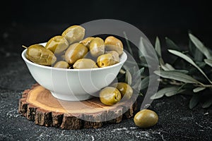 Close up shot of green olives in a white bowl on the wooden stand with olive leaves on a black background. Traditional Greek and