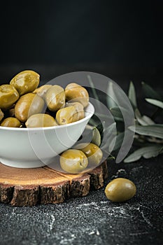 Close up shot of green olives in a white bowl on the wooden stand with olive leaves on a black background. Traditional Greek and