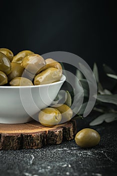 Close up shot of green olives in a white bowl on the wooden stand with olive leaves on a black background. Traditional Greek and