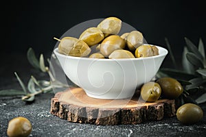 Close up shot of green olives in a white bowl on the wooden stand with olive leaves on a black background. Traditional Greek and