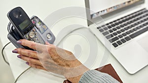 Close up shot of hand of aged woman, senior intern using phone while sitting at desk, working on laptop in modern office