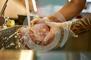 Close up shot of hands of male cook wearing protective gloves rinsing raw pork meat under tap water for cooking