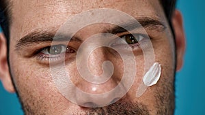 Close up shot of handsome brunette man with white cream applied on his face looking away, posing  over blue