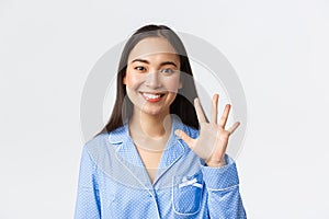 Close-up shot of happy attractive asian woman in blue pyjama showing five fingers and smiling white teeth, explain main