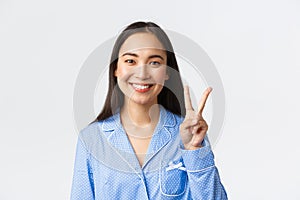 Close-up shot of happy attractive asian woman in blue pyjama showing number two and smiling white teeth, explain main