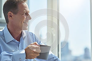 Close up shot of a happy mature businessman drinking fresh morning coffee in the office, looking in window and enjoying