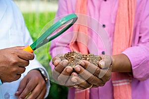 Close up shot of indian agro scientist hands checking soil from famer hand at greenhouse using magnifying glass -