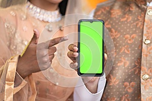Close up shot of indian muslim couple showing green screen mobile phone by pointing finger at home - concept of