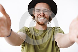 Close-up shot of lovely and happy young good-looking bearded pierced guy in glasses and hat pulling hands towards camera