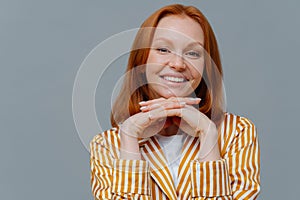 Close up shot of lovely woman has foxy hair, smiles toothily at camera, keeps hands under chin, dressed in homewear, enjoys calm