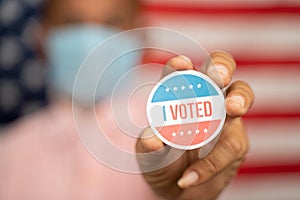 Close up shot man in medical mask showing I voted Sticker and putting on shirt with US flag as background - concept of US election