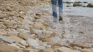 A close-up shot of a man wearing a pair of jeans and brown boots walking on the shore of a beautiful rocky beach.