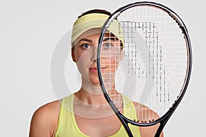 Close up shot of pleasant looking healthy woman holds tennis racket, being runner up, looks through net, wears court cap, prepares