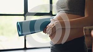 Close-up shot of pregnant belly and woman`s hand stroking tummy while other hand is holding yoga mat. Healthy lifestyle