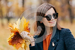 Close up shot of pretty woman with hair, wears sunglasses, has stroll during sunny day in autumn in park, carries yellow leaves, l