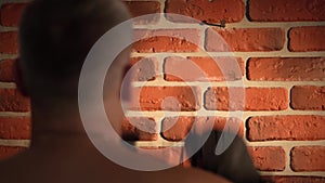 A close up shot shows a brick wall with a nail nailed to it with boxing gloves hanging from it. A man with a short