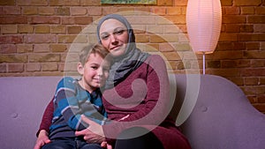 Close-up shot of small boy and his muslim mother in hijab smiling into camera caressing each other at home.