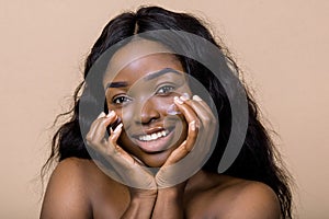 Close up shot of smiling attractive naked dark skinned woman with long hair, appiles face cream on cheeks, poses against