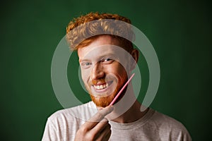 Close-up shot of smiling readhead man, combing his beard with pi