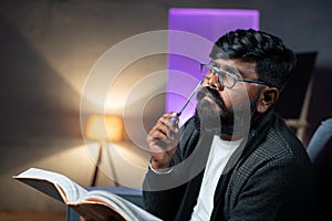 close up shot of Thoughtful Young Indian beard man while reading book during night - concept of dreaming or imagination