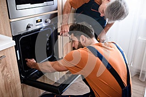 Close up shot of two repairmen, workers in uniform examining oven using screwdriver and flashlight indoors. Repair