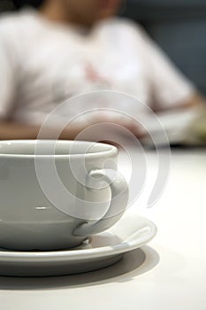 Close up shot of a white coffee cup and saucer, with an out of focus person in the background, reading a book