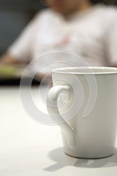 Close up shot of a white coffee mug, with an out of focus person in the background, reading a book