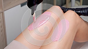 Close-up shot of woman beautician's hands in gloves applying cosmetic wax to the legs of a female client using a wooden