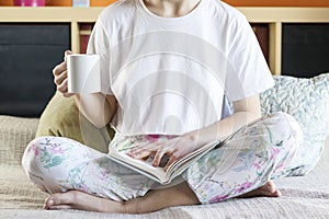 Close up shot of woman sitting in bed in morning light, by the window reading old book, having cup of cappuccino coffee
