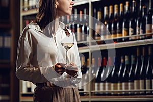 Close up shot of woman sommelier with a white wine glass stands in wine cellar with shelves full of bottles