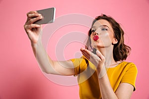 Close-up shot of young attractive woman with bright makeup sending air kiss while taking selfie on mobile phone