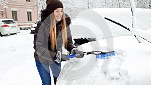 Close up shot of young happy smiling blond woman in brown coat and jeans trying to clean up snow covered car by brush