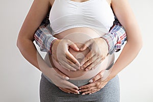 Close up shot of young man hugging his pregnant wife from behind, forming a heart shape with his hands over isolated white