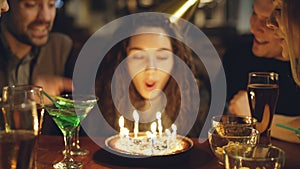 Close-up shot of young pretty woman blowing out candles on birthday cake. Happy people are laughing and clapping hands.