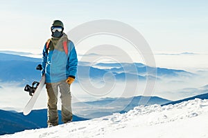 Close up shot of young snowboarder in helmet standing at the top of a mountain at golden hour with his snowboard in hand